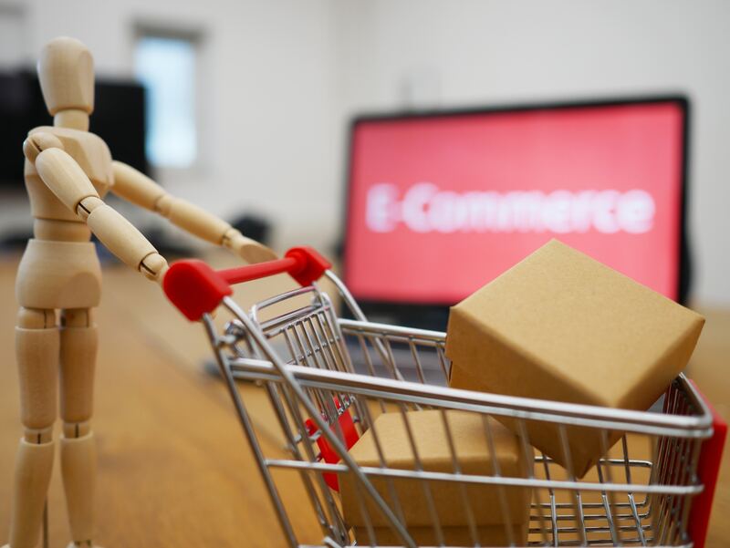 Small wooden manikin toy pushing a shopping car with packages in front of a laptop with the word e-commerce on it screen, blurred in the background